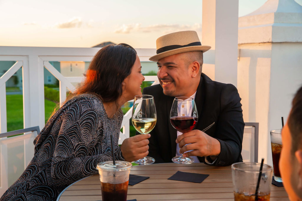 A couple smiling at each other over glasses of wine on the Encore Resort Clubhouse outdoor deck at sunset.