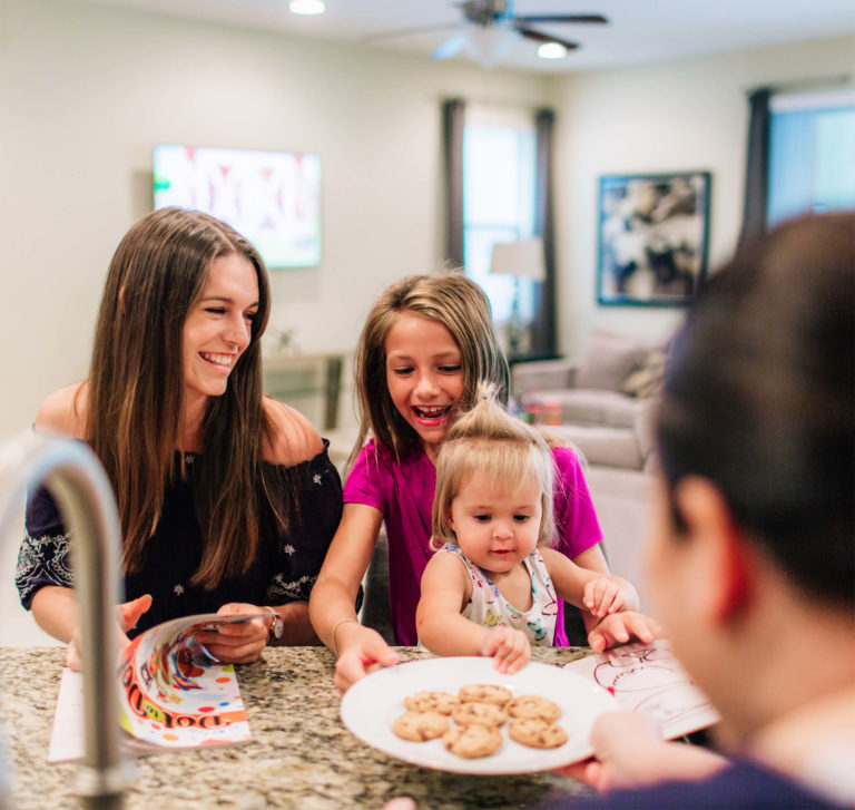 Una familia con dos hijas pequeñas comparte un plato de galletas en la cocina de un Encore Alquiler de casa de vacaciones de resort.