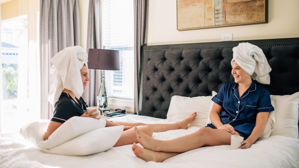 Zwei Frauen mit Badetüchern über dem Haar sitzen und unterhalten sich auf einem Kingsize-Bett in einem Encore Resort-Residenz während eines Mädchenurlaubs.