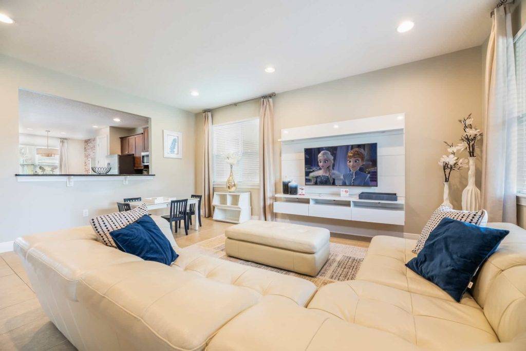 Great room with large sectional sofa, ottoman, wall-mounted TV and entertainment center, and window to kitchen