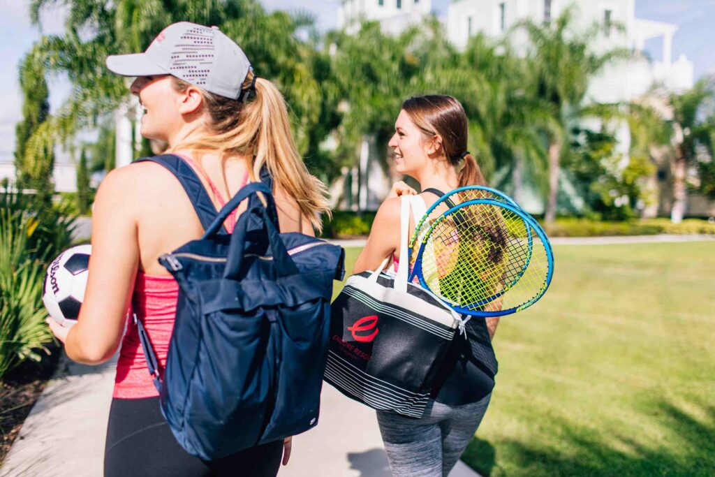 टेनिस रैकेट, खेल उपकरण के बैग और एक सॉकर बॉल ले जाने वाली दो महिलाएं Encore रिज़ॉर्ट।