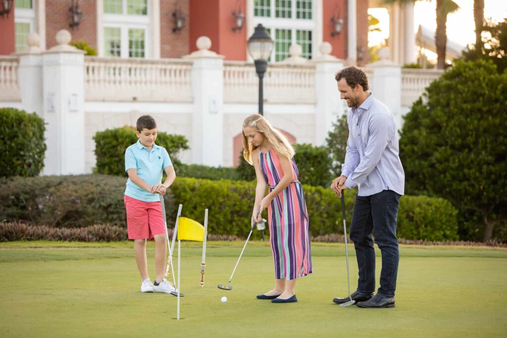 पिताजी और बच्चे हरे रंग के गोल्फ कोर्स पर गोल्फ़ खेल रहे हैं